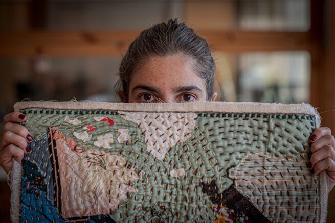 Narrativa artes textiles. Arpillera La Broma. Primer plano del trozo superior de la arpillera en manos de Gabriela Vanzetti, la creadora. Mapa realizado con trozos de telas estampadas y bordada con hilos de colores en forma de en círculos concéntricos